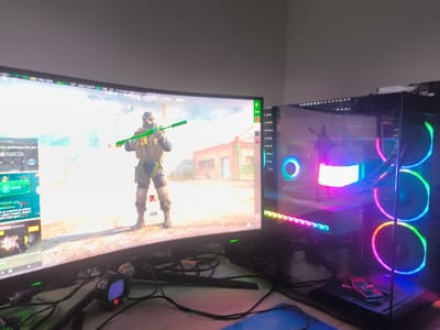 A computer monitor displays a first-person shooter video game, with a character holding a green weapon standing in an outdoor environment. To the right of the monitor is a desktop computer case with transparent side panels revealing illuminated components, including LED strips and cooling fans with color-changing lights. A digital clock reading "15:55" sits in front of the monitor amid a tangle of cables. The environment appears to be a home gaming setup. There is no location information.