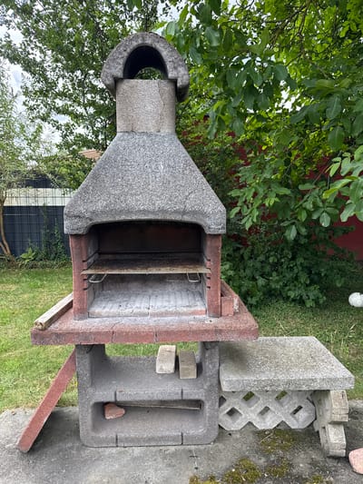 A concrete barbecue grill is situated in a grassy backyard in Weeze, Germany. The grill is multi-tiered and constructed from grey concrete with red brick accents. It features a wide hearth, two metal grilling racks, and a chimney-like structure at the top. To the right of the grill, there is a concrete bench with a decorative lattice pattern. The backyard is surrounded by lush green trees and a dark fence. In the background, a red wall is visible behind the foliage. A white soccer ball rests on the grass to the right of the bench. The sky is partly cloudy.