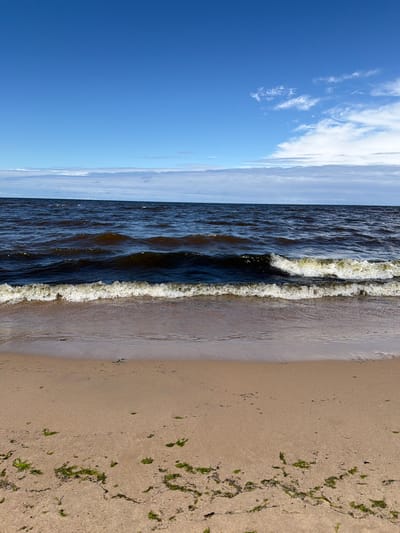This is a vertical, eye-level shot of a sandy beach in Lilaste, Latvia. The image is split into three horizontal sections: sky, sea, and beach. The sky is a vibrant blue with scattered white clouds, particularly on the right side, some of which are thin and wispy. The sea is a deep, dark blue, with small to medium-sized waves breaking on the shore. The waves have white foam crests. The beach is composed of light brown sand, with some scattered patches of green seaweed. The water meets the sand in a gentle slope, with the wet sand appearing darker and more reflective. No people or man-made objects are visible in the frame.
