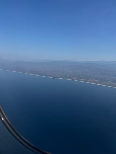 The image shows a landscape seen from an airplane window. The sky is a gradient of blue, transitioning from a darker shade to a lighter, almost hazy hue. The right side of the image is dominated by a dark blue body of water, possibly the ocean. A strip of land, likely a coastline, separates the water from a more mountainous region. The mountains appear in shades of green and brown, with visible terrain features. The edge of an airplane window is visible in the bottom right corner. The location is not discernible from this image.
