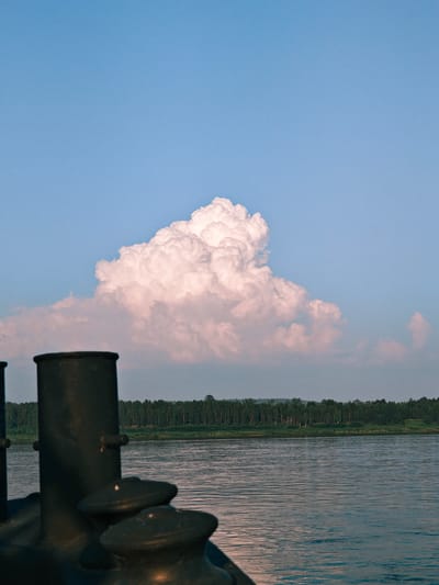 This is a photograph taken from the perspective of a boat on a wide, calm river. In the foreground, dark, metallic mooring bollards are visible. The river stretches out to a treelined bank, a dense forest of pine trees with a lighter green undergrowth. Above the treeline, the sky is a clear blue, interrupted by a large, fluffy, cumulus cloud, tinged with pink light. The scene suggests a peaceful, natural environment. The specific location is the Angara River near Lesosibirsk, Russia, a city known for its vast pine forests.