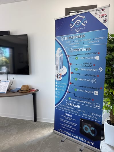 The image depicts an indoor scene, likely an office or meeting room, with a large digital display screen mounted on a white wall. To the right of the screen, a tall, retractable banner stand is prominently displayed. The banner is blue and features text and graphics related to cybersecurity. It outlines three main phases: "SE PRÉPARER" (Prepare), "PROTÉGER" (Protect), and "RÉAGIR" (React).
On a low, dark-colored table positioned in front of the screen, there is a straw fedora-style hat resting on top of some documents. A single black cable is visible on the table, connecting to something not fully in frame.
The floor is covered with a light-colored carpet, appearing to be modular tiles with a textured pattern. A portion of a potted plant is visible on the far right, in a white planter. The lighting suggests an interior environment. The banner stand is positioned to the right, obscuring a portion of the background wall, and a green plant is to the right of the banner. The overall impression is that of a business environment showcasing cybersecurity solutions.