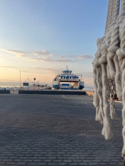 The image depicts a ferry docked at a harbor in Prinos, Greece, during what appears to be either sunrise or sunset. The sky is a soft gradient of blue and orange, with scattered clouds. In the foreground, a thick white rope hangs out of focus on the right side of the frame, obscuring a portion of the scene. A paved walkway, made of dark grey interlocking bricks, leads towards the ferry. The ferry itself is large and white with blue accents, and it is connected to the dock by ropes. Several smaller boats and a flag pole with multiple flags are visible on the dock to the left of the ferry. In the background, distant hills are silhouetted against the horizon, and the calm sea stretches out before the port. The overall atmosphere is serene and quiet, suggesting an early morning or late evening setting.