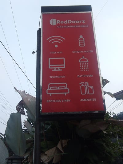 This is a medium shot, outdoor, eye-level, vertical photograph of a red sign with white text and icons, for RedDoorz hotel in Megamendung, Indonesia. The sign is mounted on a dark grey pole. The sign lists amenities such as "FREE WIFI", "MINERAL WATER", "TELEVISION", "BATHROOM", "SPOTLESS LINEN", and "AMENITIES". The sky is overcast and pale blue. There are electrical wires running across the top left of the image. To the left and bottom of the sign are large, green, tropical leaves, some of which are damaged and brown. To the right of the sign, a portion of a building with a corrugated roof is visible, partially obscured by foliage. There are no people visible in the image.