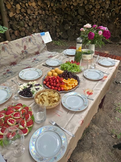 A table is set for a meal outdoors, with a rustic backdrop of stacked logs. The table is covered with a light-colored tablecloth featuring a floral pattern. On the table, there are several place settings with white plates, silverware, and glasses. A large platter holds a variety of fruits, including strawberries, apricots, cherries, and orange slices. Another dish contains cheese and olives, accompanied by a bowl of potato chips. There are also appetizers that appear to be small toasts topped with cream cheese and tomato slices. A pitcher of infused water with lemon and mint sits near a bouquet of pink and deep red peonies in a white vase. The setting suggests a casual gathering in a natural environment, possibly a garden or a picnic area. The location is identified as Буран Снт, Russia.