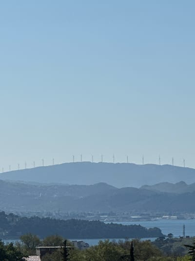 The image shows a view in Đurmani, Montenegro, featuring a mountainous landscape. Several wind turbines are lined along the crest of a mountain range. In the distance, there is a body of water adjacent to a forested area. The sky is a clear light blue. The overall scene suggests a coastal environment with renewable energy infrastructure.
