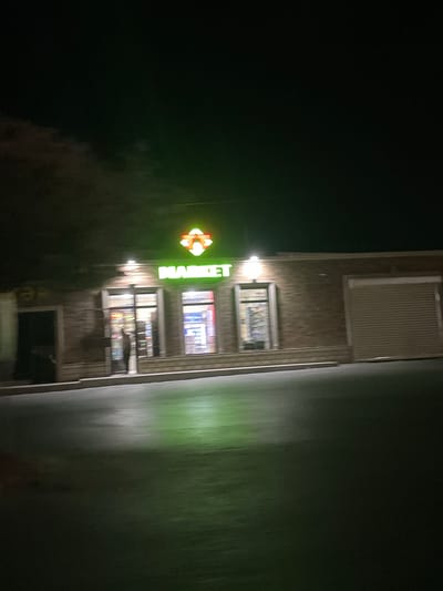 A market building with a green neon sign reading "MARKET" is visible at night. The building has a brick facade and several large windows, some of which are illuminated. A person can be seen through one of the windows. A large, closed rolling door is on the right side of the building. The area in front of the building is paved and appears wet, reflecting the green light from the sign and overhead lights. The sky is dark, suggesting it is late at night or early morning. The context places this scene in the city of Təzə Corat, Azerbaijan.