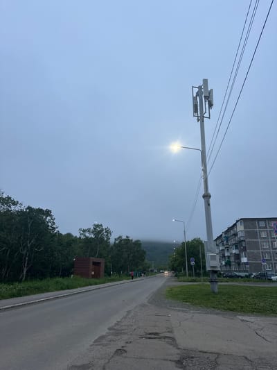 A street view captures a grey, overcast day in Vilyuchinsk, Russia. The foreground shows a cracked asphalt road with a sidewalk to the left. A line of trees borders the sidewalk, and behind them is a small, wooden, shed-like structure. Further down the road, a tall utility pole with a street light and cellular antennas is prominent. To the right of the pole, a multi-story apartment building with many windows and balconies is visible. Several cars are parked on the grass in front of the building. In the distance, a misty mountain looms. A few people are walking on the sidewalk and road, and cars are driving on the road. The overall atmosphere is subdued due to the cloudy weather.