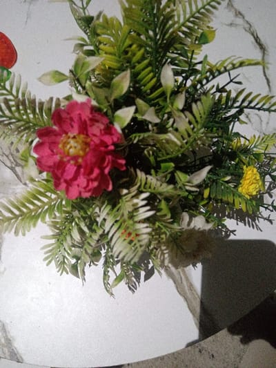 A vibrant arrangement of faux flowers and greenery sits on a white, marble-like surface. A prominent pink flower with a yellow center steals focus, surrounded by delicate fern-like foliage and smaller white and green leaves. A single yellow flower provides a contrasting accent. The composition is well-lit from above, casting a shadow in the lower right corner, suggesting the presence of a container supporting the arrangement.