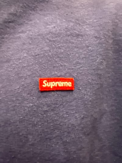 A close-up image displays a small, red, rectangular brand tag embroidered with the word "Supreme" in white capital letters. The text is oriented vertically, reading upwards from the bottom of the tag. The tag is affixed to a textured fabric surface, which appears to be dark blue or grey, possibly a garment or textile item. The fabric exhibits some subtle variations in shading and surface texture, suggesting a worn or natural fiber. There are no people, discernible activities, or specific location cues within the image, beyond the fabric and the brand tag. The setting appears to be an indoor, neutral environment focusing solely on the object.