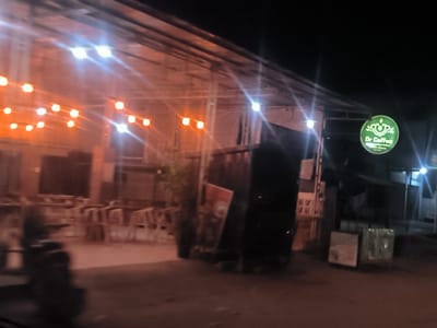 The image shows an outdoor cafe called "Dr. Coffee" in Rawang Itiek, Indonesia at night. The cafe has a metal frame structure covered with a translucent roof, decorated with strings of orange lights. Tables and chairs are visible inside the cafe. A green, circular sign with the name "Dr. Coffee" is mounted on the right side of the structure. In front of the cafe, there are dark objects that partially obscure the view.