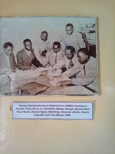 This black and white photograph, appearing to be mounted and displayed on a light-colored wall, depicts an indoor meeting of seven African men. The photograph itself is currently located in South Sakwa ward, Kenya, as specified. The event depicted occurred in Nairobi in 1958.
Six men are seated around a rectangular wooden table covered with numerous documents and papers. All seated individuals are wearing suits or collared shirts and ties, actively engaged with the papers. From right to left, the seated individuals are identified by the accompanying caption as: Chairman Oginga Odinga (rightmost, wearing a light suit, left hand on documents, looking right), Benard Mati (wearing glasses, light shirt and tie, holding a pen, looking at documents), Nzau Muimi (wearing glasses, dark suit, holding a pen, looking at documents), Masinde Muliro (wearing a dark suit and tie, holding a pen, looking at documents), and Daniel Arap Moi (far left, wearing a light suit, hands on documents, looking down). One closed, possibly leather-bound, folder rests on the table in the foreground.
Two other men are standing behind the seated group, observing the proceedings. The caption identifies Ronald Ngala as standing (prominent, wearing a light-colored shirt, looking down at the table), and Tom Mboya is presumed to be the second standing individual (partially obscured, behind Ngala and Muliro, also wearing a light-colored shirt). In the background, behind the standing figures, a stack of files or papers is visible on what appears to be a shelf or desk. The scene suggests a formal deliberation or signing event.