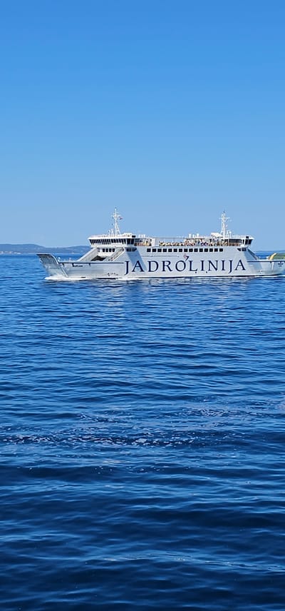 The image shows a ferry with "JADROLINIJA" written on its side, sailing on the sea. The ferry is white with blue lettering and has multiple decks with people visible on them. The sea is calm with gentle ripples. The sky is a clear, bright blue, suggesting a sunny day. In the background, a coastline with hills and vegetation can be seen. The scene appears to be set in a coastal area, possibly a harbor or a route between islands, given the presence of a ferry and the land in the distance. The overall impression is one of travel and leisure on a pleasant day. The time of day is likely daytime due to the bright blue sky and clear visibility.