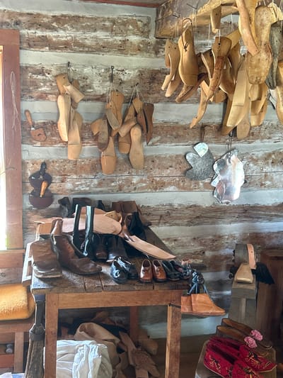 The image depicts a dimly lit room with wooden plank walls, suggesting a rustic or historical setting. The room is filled with various shoemaking tools and materials. Hung from the walls and ceiling are numerous wooden shoe lasts of different sizes and shapes, indicative of a shoemaker's workshop. On a wooden table in the foreground, there are several pairs of boots and shoes, some appearing old and worn, others newer and made of leather. Various leather scraps and shoe components are also visible on the table and floor. In the background, a large wooden workbench with tools, possibly a shoe press or other equipment, is present. The overall impression is that of a preserved or active shoemaking studio. The provided metadata indicates this scene is located in South Dundas, Canada.
