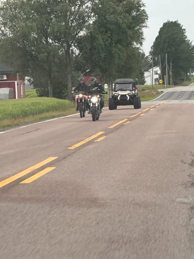 Two individuals on motorcycles are traveling on a paved road with double yellow lines in the foreground. A white all-terrain vehicle is also on the road behind the motorcycles. In the background, there are trees and a red building with a white roof. A white house with a brown roof is visible further down the road, along with a speed limit sign indicating "60". The environment appears to be rural.