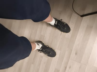 The image shows a low-angle view of a person's legs and feet, wearing dark blue pants and white athletic socks with black Nike shoes. The shoes are primarily black with a neon green swoosh logo and accents. The person is standing on a light-colored wooden floor. In the upper right portion of the image, a black cord, possibly from an electronic device, is visible on the floor. The setting appears to be indoors, given the flooring and the presence of the cord. There is no indication of the time of day or weather conditions, and no discernible emotions are conveyed. The text "NIKE" is visible on the side of the shoes, and a small Nike swoosh is also present on the toe area of both shoes. The location context is Troisdorf, Germany.