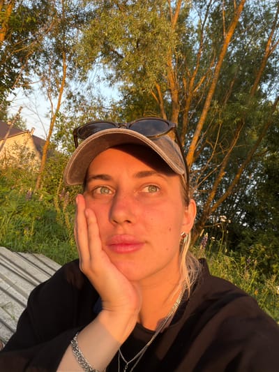 This is a close-up, portrait-style photograph of a young person lying down outdoors. The person is wearing a grey baseball cap, sunglasses perched on top, and a black top with a silver chain necklace. Their left hand is resting on their cheek, and their expression is relaxed, with their lips slightly pursed. The sun is setting, casting a warm, golden light on the scene, illuminating the person's skin and the surrounding foliage.
The background consists of lush green grass and trees with visible branches. In the distance, a small house with a light-colored facade and a red roof can be seen. The overall atmosphere is peaceful and serene, suggesting a casual moment of relaxation in nature. The time of day is clearly sunset, indicated by the warm, low-angled light. The location cues, combined with the user-provided context "Пропан Снт, Russia," suggest a dacha or countryside setting in Russia. There is no visible text in the image.