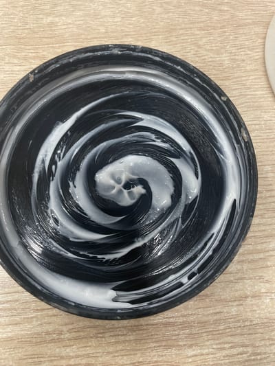 An overhead view of a black jar lid, with white cream swirled in a spiral pattern on its surface. The lid is resting on a light brown, wood-grain surface. The scene is indoors. This image is not relevant to the city of Терволово, Russia.