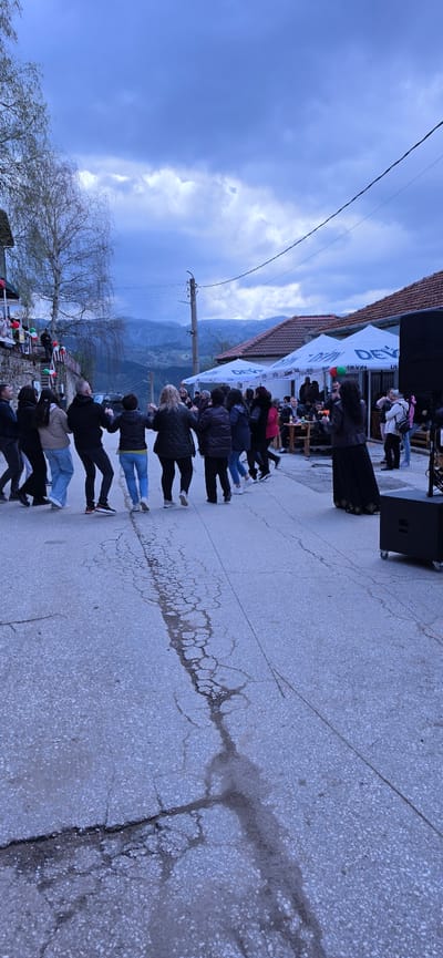 The image shows a group of people lined up and potentially dancing in Breze, Bulgaria. They are on an asphalt street with visible cracks. A stage or structure is set up to the right of the frame, with a speaker and possibly a performer. In the background, there is a building with a roof and a "DEVIN" branded tent. Mountains are visible in the distance. The sky is overcast. Power lines run along the left side of the frame.