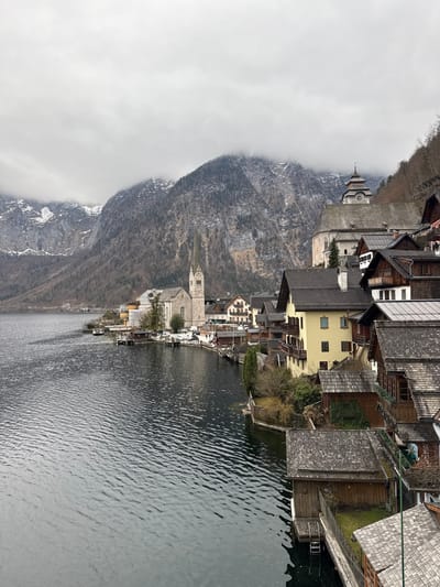 This elevated view captures the village of Hallstatt, Austria, nestled along the western shore of the Hallstätter See, with steep, snow-dusted mountains rising immediately behind it under an overcast sky. The dark, rippled surface of the lake dominates the left and lower-center foreground.

Along the lakefront, a dense collection of traditional multi-story buildings with prominent gabled roofs, primarily dark grey or brown, extend up the hillside. The Hallstatt Lutheran Church, a light-colored stone structure with a tall, slender spired clock tower, stands near the water's edge on the left side of the village. Higher on the hillside, behind and to the right of the Lutheran church, the Catholic Parish Church of the Assumption is visible, distinguished by its unique multi-tiered, helmet-shaped tower roof.

Residential and commercial structures, some painted light yellow, others in natural wood tones or white plaster, are built into the steep terrain, often with balconies and foundations extending to the lake. Multiple small, wooden boathouses are constructed directly over the water or at its edge. A narrow road or pathway runs along the lakefront, with at least one white passenger vehicle parked near the Lutheran church. A small white boat is moored at a dock near the church.

The background features large, steep mountains heavily forested with bare deciduous and some evergreen trees. Their upper slopes display exposed rock and patches of white snow, partially obscured by low-hanging grey clouds. No individuals, explicit actions, or interactions are visible within the frame. The scene presents a static, panoramic view of the village and its natural environment.