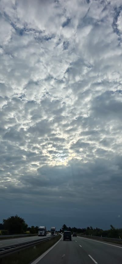 The image shows a view from a vehicle traveling on a highway in Vejen, Denmark. The sky is predominantly filled with clouds, with patches of blue sky visible. The sun is partially obscured by clouds, casting rays of light downwards. On the highway, several vehicles are visible, including two trucks in the left lane and a black car in the right lane. The road has multiple lanes, separated by white lines. The sides of the highway are lined with trees and vegetation. The overall scene depicts a cloudy day on a highway in a rural or semi-rural area.