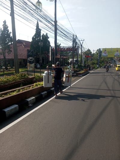 A man walks along the side of a road, carrying two large white containers suspended from a pole across his shoulders. He is wearing a dark shirt, dark pants, a baseball cap, and sandals. The road is asphalt, with a white dividing line and a curb painted with black and white stripes. To his left is a raised garden area with a building in the background. Overhead are numerous electrical wires. Further down the road, there is a billboard advertising "Selamat Hari Raya Idul Fitri". A motorcycle and a yellow vehicle are also visible on the road. Based on the environment, including the writing on the billboard, this image is not from Nhot Chang, Laos.