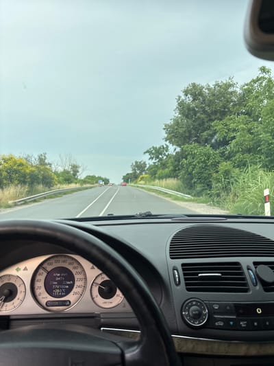 The image is taken from the perspective of the driver of a Mercedes-Benz vehicle, looking forward through the windshield. The car is on a paved road with two lanes in each direction, separated by white lines. The road is bordered by greenery, with trees and bushes on either side. The sky is overcast.
In the foreground, the steering wheel and a portion of the dashboard are visible, including the instrument cluster. The instrument cluster displays the following information:
- Speedometer: indicated speed is between 0 and 40 km/h, with the needle pointing around 20 km/h.
- Tachometer: needle is between 0 and 1,000 rpm, pointing at approximately 800 rpm.
- Digital display:
- "After start": 0:47 h
- 25 km
- 7.7 L/100km
- +28.0 °C
- 111.4 km
- Odometer: 432832 km
In the midground, several cars are visible on the road ahead, traveling in the same direction. There is a guardrail on the left side of the road, and a white and red marker post on the right.
The image was taken in or near Yoakim Gruevo, Bulgaria, as indicated by the metadata. The environment appears to be rural or semi-rural, with vegetation suggesting a natural landscape. The lighting suggests it is daytime, but the overcast sky reduces the intensity of sunlight. There are no people clearly visible in the image. The overall scene depicts a car journey on a road in Bulgaria.