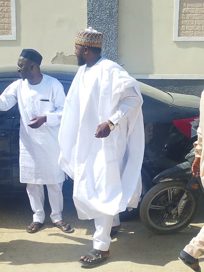 The image depicts an outdoor scene in Darmanawa, Nigeria, during daylight hours. Two adult males are central to the frame, with partial views of a third individual and several vehicles.
In the center, a man with dark skin and a beard stands facing right, wearing a white traditional kaftan-style garment over white trousers and a multi-colored, patterned cap (kufi). He wears a gold-colored wristwatch on his left wrist and dark sandals with a light-colored chevron design. His left hand is slightly raised.
To the left, a second man with dark skin stands facing left, gesturing with open hands. He wears a plain black cap, a long-sleeved white traditional tunic with a small dark pocket on the left chest, white trousers, and dark sandals.
Behind these two individuals, a dark-colored sedan is partially visible, parked or stationary, showing a portion of its rear and a red-and-white tail light assembly. To the right of the sedan, a black motorcycle's front wheel, tire, and suspension are partially visible.
In the background, a light-colored building with textured architectural details, including rectangular decorative frames and raised patterns, is visible. The ground is unpaved, appearing as compacted dirt or sand. Shadows suggest direct sunlight. A partial view of a third person's lower legs and feet, wearing light-colored trousers and dark sandals, is visible in the lower right foreground.