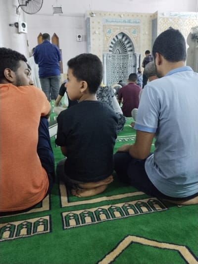 The image shows a mosque interior with a green carpet. Four men are sitting on the carpet facing the prayer niche (mihrab), which is decorated with intricate mosaic patterns and a large archway. The youngest of the men is sitting on his legs with his feet tucked underneath him. He has short black hair and is wearing a black shirt. The man to his right is also sitting on his legs and has short black hair. He is wearing a blue shirt and dark blue pants. The man sitting to the left of the youngest man is also wearing a blue shirt and pants. He is sitting with his legs crossed. The man to his left is older with short black hair and a beard. He is wearing an orange shirt. The men are all looking towards the mihrab. The image is taken from a low angle.  This scene appears to be in a mosque in the New Bani Sewif City, Egypt.  The coordinates are (29.027, 31.112).