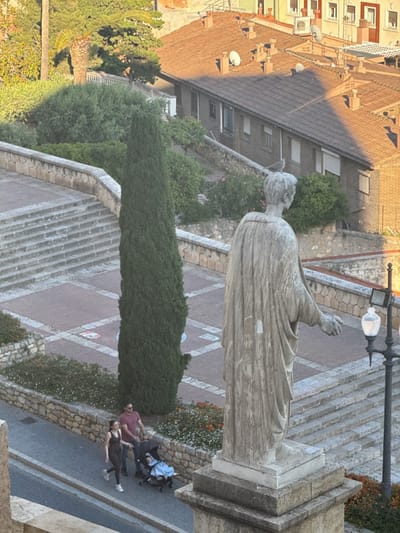 The image shows an elevated view of a scene in Tarragona, Spain. A light-colored stone statue stands prominently in the foreground, facing away from the viewer. The statue is positioned on a multi-tiered stone base.
A couple walks along a sidewalk, pushing a stroller.
Behind the statue, a tall, thin cypress tree rises among other greenery. The background includes a series of stepped walkways, tiled areas, and multi-story buildings with reddish-brown tile roofs. The environment is bathed in warm, late-afternoon or early-morning sunlight.