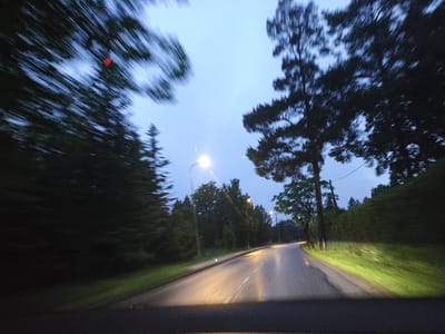 The image depicts a blurred view from a moving vehicle on a road at dusk or dawn. The road is paved, wet, and illuminated by headlights and streetlights. Tall trees line both sides of the road, with their foliage appearing dark and indistinct due to motion blur. The sky is a pale, overcast blue. In the distance, there are more trees and possibly some buildings, though they are not clearly discernible. The location is identified as Harkujärve, Estonia. There are no people or clear subjects visible in the frame. The overall impression is one of travel through a wooded area during low light conditions.