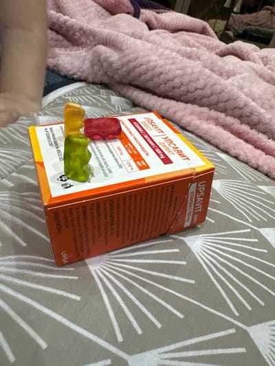 Here's a detailed description of the image:
**People:** Only a small portion of a person's arm and hand are visible at the bottom left of the image.
**Subjects:** The primary subjects are a box of "UPSAVIT DIRECT" Vitamin C 1000mg and three gummy bear candies.
**Objects:** The image includes a box of Vitamin C supplements, three gummy bear candies in red, yellow, and green, and the background features a patterned grey and white blanket and a pink fuzzy throw or blanket.
**Actions/Interactions:** The gummy bears are resting on top of the Vitamin C box. The box is unopened, secured with a rubber band.
**Environment:** The photo seems to have been taken indoors, possibly on a bed or couch, based on the blankets in the background. The precise location is not determinable from the image, and no specific city or place is identifiable. The package, however, indicates it has been notified to the Ministry of Health. The mention of a tel number (+40372582288) suggests that it may be associated with either Bucharest, Romania as the +40 area code is commonly associated with Romania.
**Details from the Packaging (OCR):**
* The box contains Vitamin C 1000mg.
* It notes how much Vitamin C per daily serving.
* It lists some of the ingredients.
* The side of the box has details about the manufacturer, producer, distributor, and a net weight of 54g.
* It indicates the package is recyclable, displaying a PAP symbol.
* It mentions it is notified to the Ministry of Health.