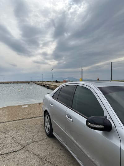 This image captures a scene in Limenas Avdiron, Greece, under an overcast sky. The foreground and midground on the right display the passenger side of a silver four-door Mercedes-Benz E-Class sedan, parked on cracked concrete pavement. The vehicle exhibits tinted windows and a black side mirror equipped with integrated LED turn signals. To the left, a concrete pier extends into a calm body of water, where several red and white buoys are visible floating. Approximately 3-4 indistinct human figures are standing on the pier in the mid-distance. Multiple utility poles or streetlights are positioned along the pier and the adjacent shoreline. In the background, distant landmasses are discernible across the water, situated beneath the extensive gray cloud cover.