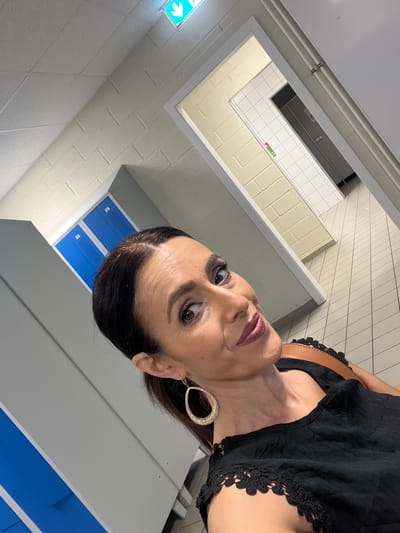 In Mensdorf, Luxembourg, a woman with dark hair, styled back and pulled into a ponytail, takes a selfie. She is wearing a black top with intricate lace trim on the sleeves and neckline. Large, circular, rhinestone-encrusted earrings adorn her ears. A brown strap, likely from a purse, is visible over her shoulder. The background appears to be a locker room. Bright blue lockers are stacked to the left, and to the right, a doorway reveals a tiled hallway with more lockers. The ceiling features a fluorescent light with a green exit sign. The floor is tiled with light-colored square tiles. The woman's gaze is directed towards the camera with a slight smile and berry-colored lipstick.