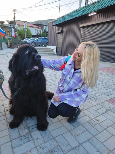 A full-shot captures a woman and a large, black Newfoundland dog on a paved walkway in Орджоникидзевский поселковый совет, Ukraine. The woman, with shoulder-length blonde hair, is crouching slightly. She wears a light purple and white checkered flannel shirt, a black top underneath, black leggings, and dark shoes. She is petting the dog with her right hand, smiling. The Newfoundland is sitting, attached to a leash. The dog's fur appears wet and somewhat matted. In the background, there is a building with brown corrugated metal siding and a green, overhanging roof. To the left, there's a partial view of a street with parked cars, buildings, and flagpoles flying the flags of Russia and what appears to be France. Power lines are visible in the sky. The pavement is composed of interlocking rectangular pavers.