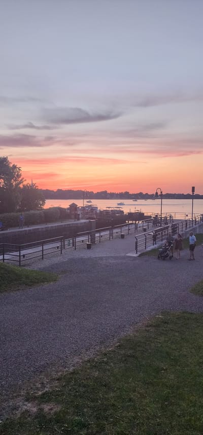 The image captures a sunset over the Chambly Canal in Chambly, Canada. The sky displays a gradient of soft pinks and oranges, transitioning into pale blue with scattered clouds. The canal, a prominent feature, is flanked by dark, weathered stone walls and bordered by metal railings. A paved pathway runs alongside the canal, with gravel paths branching off into grassy areas.
Several boats are visible on the water, anchored or moving slowly. On the pathway, a family is walking. A man, woman, and child in a stroller are moving together, with another man standing nearby. Further in the background, a lone figure stands on the canal's edge, silhouetted against the twilight sky. Streetlights with ornate poles are positioned along the pathway. The overall scene suggests a tranquil evening at a popular recreational spot.