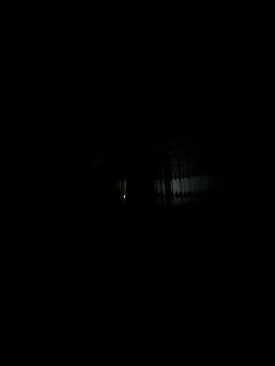 The image is a nighttime or very dark scene. The primary subject appears to be a body of water, possibly a canal or river, reflecting some form of light source. The water's surface is disturbed, creating rippling patterns of light and shadow. In the upper part of the image, the reflection suggests a structure with horizontal elements, possibly a bridge, railing, or even a building with distinct lines. There is a faint, rectangular light source visible in the central area of the image, illuminating the disturbed water. The overall impression is one of darkness and mystery, with the light on the water being the only significant focal point. There is no visible text. The scene likely takes place outdoors at night, given the lack of ambient light and the water's reflection of a light source. The location context of Paytugh, Uzbekistan, does not provide specific visual cues within the image itself.