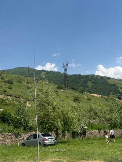 The image depicts a sunlit outdoor scene in Aghveran, Armenia. A silver sedan is parked on the left side of the frame, angled into the grassy area. In the mid-ground, two individuals, a woman and a girl, walk across the lawn away from the viewer. The background features a lush, green, tree-covered hillside under a clear blue sky dotted with white clouds. A tall, metal electricity pylon stands on the hillside, with power lines stretching across the frame. The foreground is characterized by a stone retaining wall bordering a grassy area with scattered trees and vegetation. The overall impression is one of a natural, rural landscape on a bright, clear day.