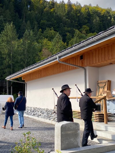 The image depicts an outdoor scene in Abondance, France, featuring a modern building against a backdrop of a dense, green forest covering a steep hillside under an overcast sky. The single-story building has a white facade, a dark tiled roof with light wooden eaves and soffit, and a grey metal gutter system. The ground in front of the building is covered with grey gravel, and a concrete barrier is positioned in the foreground.
Four individuals are present. On the right, two men are dressed in dark jackets, trousers, and dark hats. One man, with glasses and a beard, is observed in left profile. The second man is ascending concrete steps towards the building's entrance, his back largely to the viewer. Two bagpipes, featuring dark bags and light-colored chanters and drones, are propped vertically against the building wall near these men.
To the left of the frame, a woman with reddish-brown hair, wearing a dark jacket, blue jeans, and brown shoes, walks away from the camera. Adjacent to her, a man in a dark jacket, light blue jeans, and dark shoes also walks away, with a strap visible over his shoulder. A white commercial van is partially visible behind them on the far left. Stacked near the building's entrance are multiple wooden folding tables/benches, several cardboard boxes, and a white container.