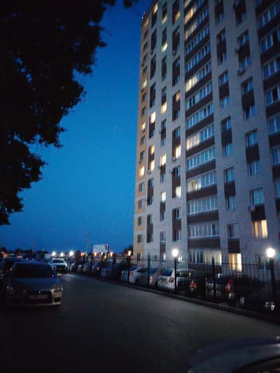The image depicts a nighttime scene in the city of Krutye Vyselki, Russia, focused on a tall, multi-story apartment building. The building, constructed with light-colored brick and dark brown architectural elements, features numerous windows, many of which are illuminated, suggesting occupancy. A large tree with dark foliage occupies the left side of the frame, partially obscuring the building and the twilight sky, which is a deep blue. In the foreground, a paved area, likely a street or parking lot, is visible with several cars parked. A dark-colored sedan is prominently positioned in the lower left, displaying a license plate. A dark metal fence runs along the front of the apartment building, separating it from the parking area, and several lampposts with bright lights are interspersed along the fence. The overall atmosphere is urban and residential, captured during the evening or early night.