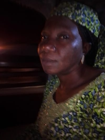 A woman with dark skin is pictured from the chest up, looking slightly to her right. She is wearing a headscarf and a green and blue patterned dress adorned with small, shimmering embellishments around the neckline. Her right ear is visible and she is wearing a small, light-colored earring. The background is dark and blurred, with horizontal lines suggesting wooden surfaces. The lighting is dim and appears to be artificial. The image is consistent with a candid photograph taken indoors in a dimly lit environment. There is no information in the image to determine the specific location within Nassarawo, Nigeria.