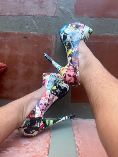 A pair of high-heeled platform pumps, adorned with a glossy, multi-colored collage print of magazine covers and pages featuring various models and prominent text such as "VOGUE" and "EZEXZ," are being worn on two light-toned, visibly hairy human feet/lower legs. The shoes feature thin stiletto heels with black tips and thick platforms. The feet and shoes are positioned on a horizontal surface composed of reddish-brown square tiles separated by grey grout lines. A dark grey-brown vertical surface forms the background. A corner of a textured orange object, possibly fabric or a cushion, is visible in the lower-left foreground. The scene is situated in Arraial Do Cabo, Brazil.