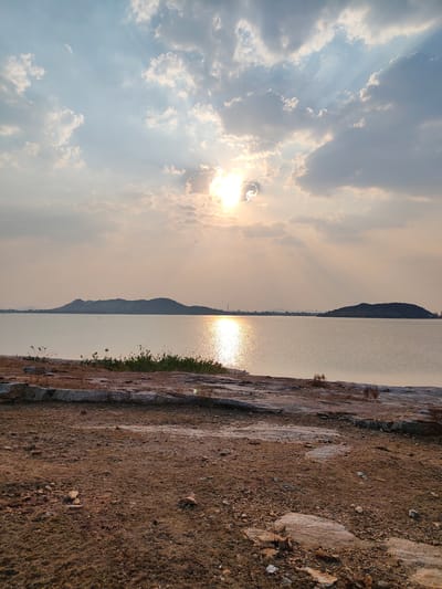 The image depicts a tranquil scene at a lake in Kammavaripalli, India, under a sky filled with clouds partially obscuring the sun. The sun's rays stream down, creating a shimmering reflection on the water's surface. Two landmasses appear in the distance. The foreground features dry, rocky terrain leading to the water's edge. Sparse vegetation is visible near the waterline. There are no people visible.