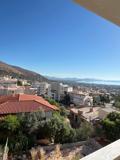 A bright, clear, sunny day over a coastal town with a body of water in the distance. The scene captures a panoramic view from a slightly elevated position, looking down and out over a residential area. In the foreground, there is a terracotta-tiled roof of a house with green trees and shrubs surrounding it. Further into the midground, a densely populated urban landscape unfolds, characterized by multi-story buildings with white and cream facades. Some buildings appear to be modern, while others show signs of older architecture, indicated by the presence of red-tiled roofs. The terrain rises behind the town, forming a barren, rocky hill covered in sparse vegetation. In the background, a vast expanse of water, likely a sea or large bay, is visible, with mountains or hills forming a distant horizon. The sky is a clear, vibrant blue with no clouds. The overall impression is of a Mediterranean coastal town on a warm, sunny day.