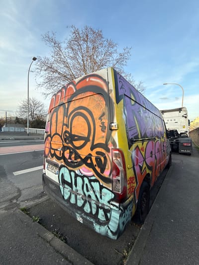 This image captures a vibrant, graffiti-covered van parked on the side of a road during what appears to be daytime. The perspective is from slightly above and to the rear of the van, looking towards its front and side. The sky is clear and blue with minimal clouds, suggesting fair weather.
The primary subject is a white utility van, completely adorned with colorful graffiti art. The graffiti features bold, interconnected letterforms and abstract shapes in various bright colors including orange, pink, purple, yellow, light blue, and black outlines. On the rear of the van, visible across the back doors, is a large orange and black piece, while the side of the van is covered in purple and yellow designs. The van's rear lights are visible, indicating the back of the vehicle. A European-style license plate is mounted on the lower left rear door, partially obscured by graffiti, but "0D-454-ZZ" is clearly visible. Below the right taillight, the brand name "RENAULT" is visible in embossed letters. The van appears to be parked close to a curb, with its right rear wheel almost touching the edge of the paved road or sidewalk.
In the background, behind the van and further down the road, a large white semi-truck is parked. The rear of its cab and a portion of its trailer are visible. On the back of the truck's cab, the brand name "MAN" is visible, along with numbers like "68 80 90".
To the left of the van, in the middle ground, there's a deciduous tree with bare branches, suggesting it might be late autumn or winter. Further in the distance, another similar tree can be seen. A street light with a curved pole is also visible above the van and to its left. The road surface where the van is parked is asphalt, and there are some white road markings. A concrete barrier or wall runs along the side of the road further back.
