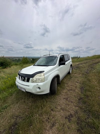 A white Nissan X-Trail SUV is parked on a grassy hill with a dirt track in front of it. The car is facing the right side of the image. The license plate reads "BY 8662 IC". The background shows a landscape with trees, fields, and a cloudy sky. The location is Lysohirka, Ukraine.
