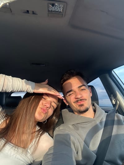 A man and woman are posing for a selfie inside a car. The man has dark hair and a beard, wearing a gray hoodie and a seatbelt. The woman has long brown hair, wearing a white top, and is making a kissing face while holding her hand up to her forehead. The car's interior is gray. The location is unknown, and there is no evidence to suggest the city of Chokoba, Bulgaria.
