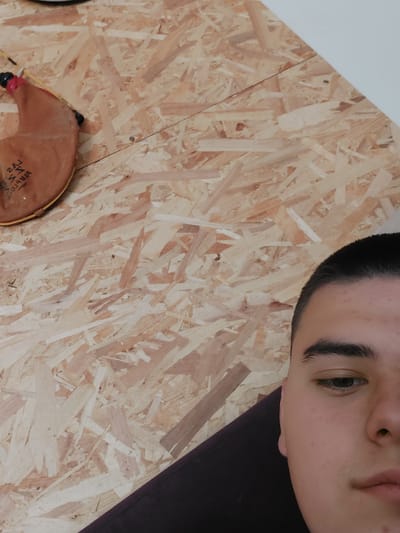 A close-up, diagonally oriented shot shows a young man's face in the upper right of the frame, set against a background of oriented strand board (OSB). The man has dark hair and a visible eyebrow. In the lower left corner, a brown, circular object with writing on it is partially visible. The background OSB appears to be part of a wall or surface. The location of Novo Zhelezare, Bulgaria is inferred from the source.
