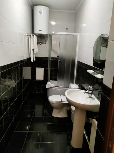 This is a forensic, matter-of-fact description of a bathroom located in San Martín de Porres, Peru.
The scene depicts a small, brightly lit bathroom with black and white tiled walls and black tiled flooring. In the foreground, a white pedestal sink with a chrome faucet is visible. To the left of the sink is a white toilet with a blue and white sticker on the seat. Behind the toilet, a glass-enclosed shower stall with frosted glass doors is situated.
Mounted on the wall above the shower stall is a white cylindrical water heater. To the left of the water heater, a chrome towel rack holds several folded white towels. In the upper right corner, a circular mirror is mounted on the wall, reflecting a portion of the bathroom.
On a clear glass shelf above the sink, two small white bars, possibly soap, are placed. To the right of the sink, a white rectangular object, likely a radiator or ventilation unit, is positioned vertically against the wall. The overall environment is clean and well-maintained, suggesting a hotel bathroom or a similar lodging facility. There are no people or any signs of recent activity in the image.