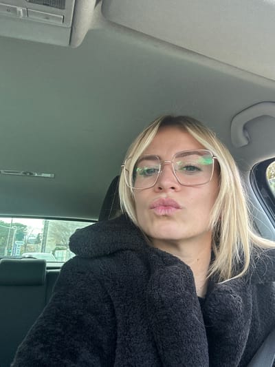 A blonde female is depicted from the upper chest upwards, seated within a vehicle. She wears square-framed, thin-rimmed spectacles that reflect green and white light sources. Her hair is blonde, parted on the side, and frames her face. She is dressed in a dark, textured garment, resembling fleece or faux fur. Her lips are pursed into a 'kiss' expression.

The vehicle's interior features a light grey headliner. A black headrest is partially visible behind her left shoulder, and a light-colored grab handle is visible on the right.

Through the vehicle's rear window, an exterior street scene in Vallauris, France, is visible. A prominent green street sign with white lettering on the left displays "PARKING PAYANT 2H" and "PHARMACIE". Beyond the sign, a light-colored vehicle is partially visible. Deciduous trees with sparse branches suggest a non-summer season. A building structure is partially visible behind the trees. The sky above is clear. The subject is looking directly towards the camera.